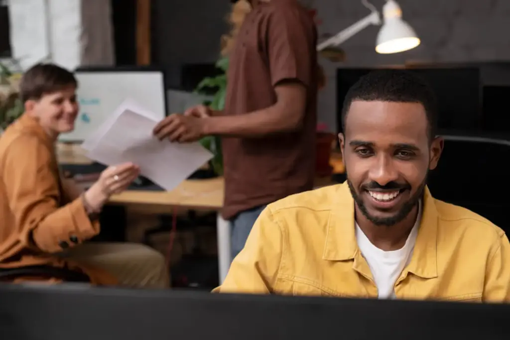 Rapaz sorrindo em frente ao computador com colegas ao fundo conversando.