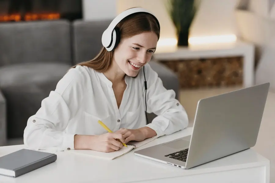 Jovem mulher sorri para a tela do seu notebook enquanto faz anotações de uma aula, representando Google Classroom.