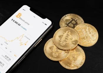 Moedas físicas de Bitcoin ao lado de um celular com gráficos de criptoativos na tela, representando comprar criptomoedas no Brasil.