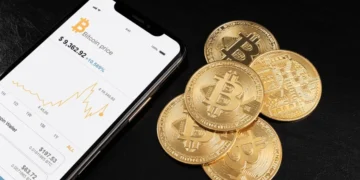 Passo a passo para comprar e vender criptomoedas no Brasil 5 Moedas físicas de Bitcoin ao lado de um celular com gráficos de criptoativos na tela, representando comprar criptomoedas no Brasil.