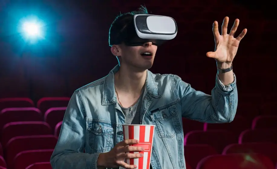 Homem usa óculos de realidade virtual em sala de cinema, enquanto segura um copo de pipoca, representando inovação no entretenimento.