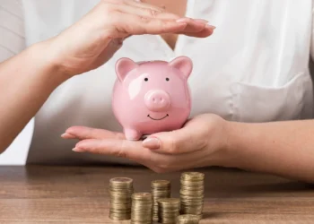 Mulher segura cofre em formato de porco. Há pilhas de moedas sobre a mesa. Falando de investir em renda fixa.