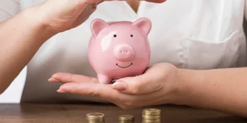 Mulher segura cofre em formato de porco. Há pilhas de moedas sobre a mesa. Falando de investir em renda fixa.