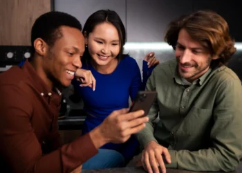 Três amigos animados olhando um iPhone 16 juntos em um ambiente aconchegante.