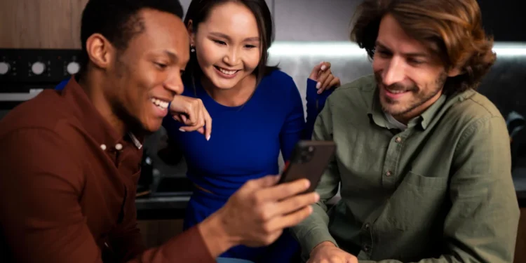 Três amigos animados olhando um iPhone 16 juntos em um ambiente aconchegante.