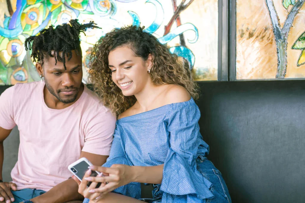 Duas pessoas sorrindo enquanto olham um iPhone 16 juntas em um ambiente colorido.