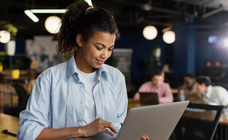 Jovem aprendiz trabalha em um computador. Há colegas de trabalho no fundo da imagem.