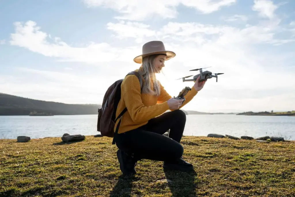 Mulher ajoelhada em gramado, segurando drone ao lado de um lago sob luz do dia.