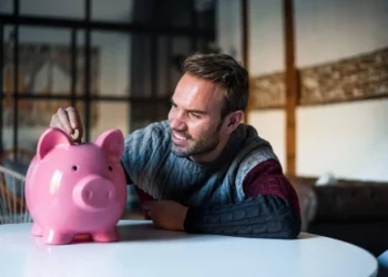Homem sorridente poupando dinheiro em cofrinho, simbolizando investimentos para viver de renda.