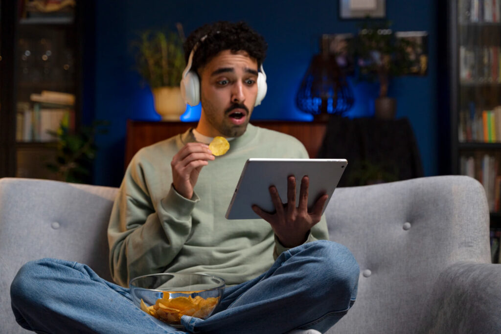 Homem surpreso assistindo Paramount+ no tablet, com fone de ouvido e tigela de batatas na sala.