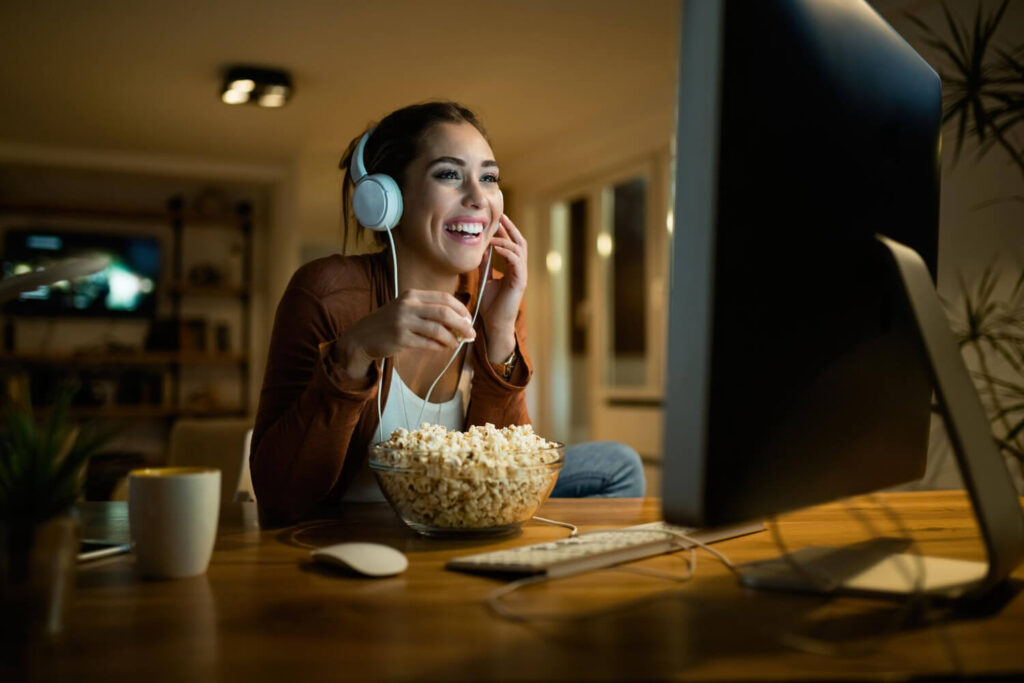 Mulher sorrindo, assistindo Paramount+ no computador, com fone de ouvido e tigela de pipoca na mesa.