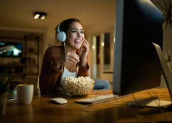 Mulher sorrindo, assistindo Paramount+ no computador, com fone de ouvido e tigela de pipoca na mesa.