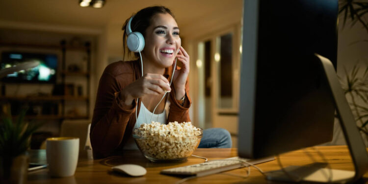 Mulher sorrindo, assistindo Paramount+ no computador, com fone de ouvido e tigela de pipoca na mesa.