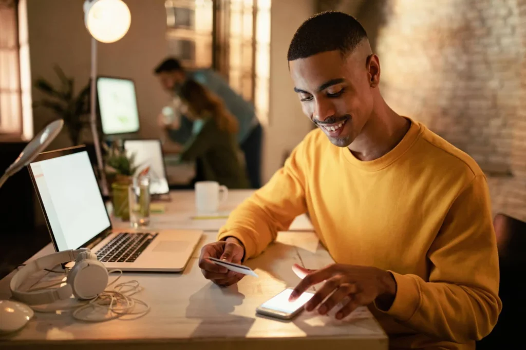 Homem sorrindo enquanto usa o celular para acessar o Bom Pra Crédito, segurando um cartão na mão.