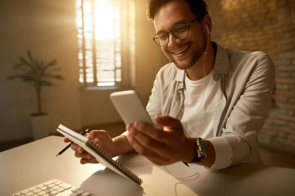 Homem sorrindo enquanto pesquisa sobre Bom Pra Crédito no celular, com caderno e fones de ouvido.