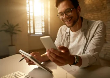 Homem sorrindo enquanto pesquisa sobre Bom Pra Crédito no celular, com caderno e fones de ouvido.