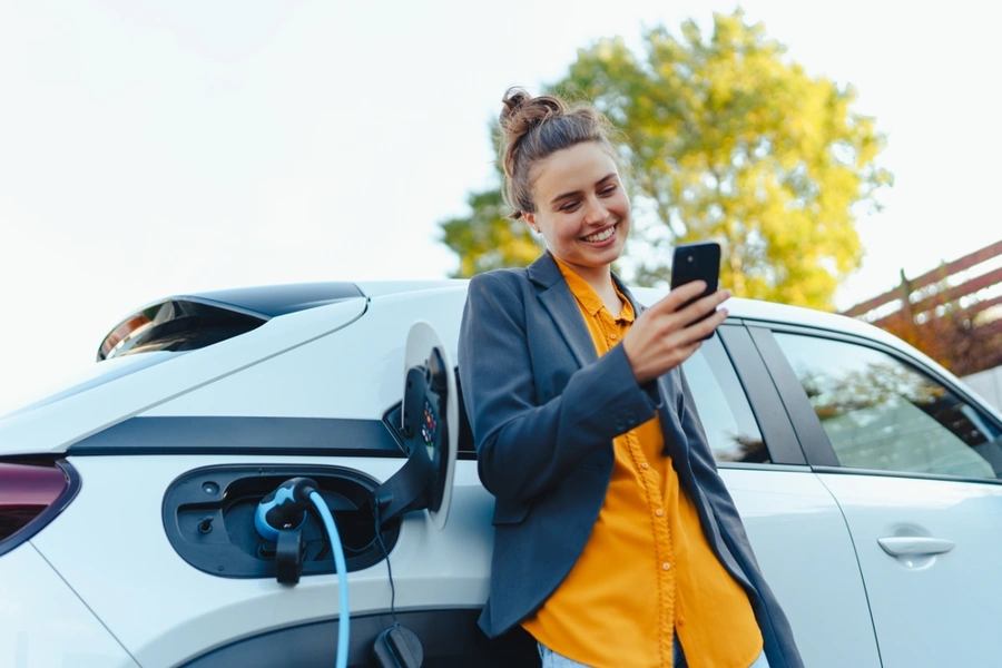 SUVs e carros elétricos: como será a mobilidade no Brasil em 2025? 17 Condutora sorridente mexe no smartphone enquanto seu suv elétrico recebe uma recarga. Falando sobre a mobilidade no Brasil.