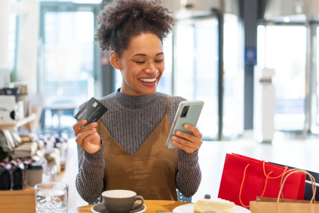 Mulher sorrindo em cafeteria, segurando celular e cartão, representando cartões de crédito sem anuidade.