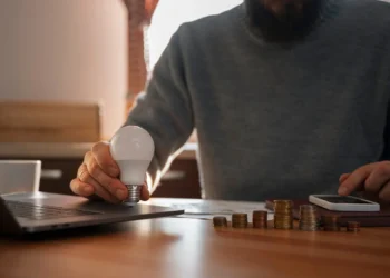 Homem segura lâmpada próximo a uma calculadora, uma pilha de moedas e um notebook. Falando de conta de luz.
