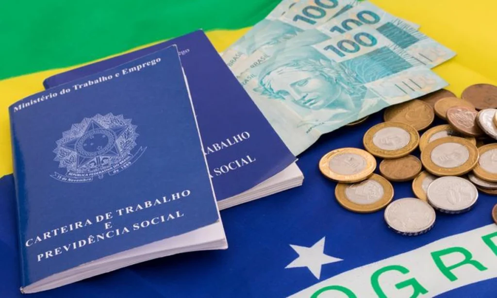 Duas carteiras de trabalho sobre uma bandeira do Brasil com algumas moedas e notas de R$ 100 ao lado. Falando de empréstimo consignado CLT.