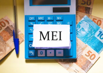 Bloco de notas escrito "MEI". Ele está sobre uma calculadora e algumas cédulas espalhadas numa mesa.