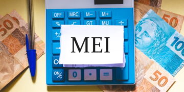 Bloco de notas escrito "MEI". Ele está sobre uma calculadora e algumas cédulas espalhadas numa mesa.