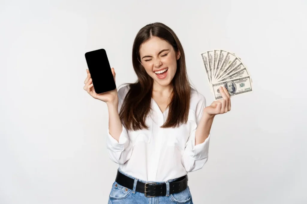 Mulher sorri enquanto exibe um celular numa mão e um maço de cédulas de dólar na outra. Representando como ganhar dinheiro na internet.