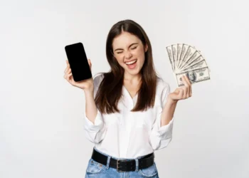 Mulher sorri enquanto exibe um celular numa mão e um maço de cédulas de dólar na outra. Representando como ganhar dinheiro na internet.