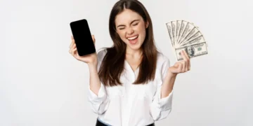 Renda extra online: 10 maneiras seguras de ganhar dinheiro na internet 3 Mulher sorri enquanto exibe um celular numa mão e um maço de cédulas de dólar na outra. Representando como ganhar dinheiro na internet.