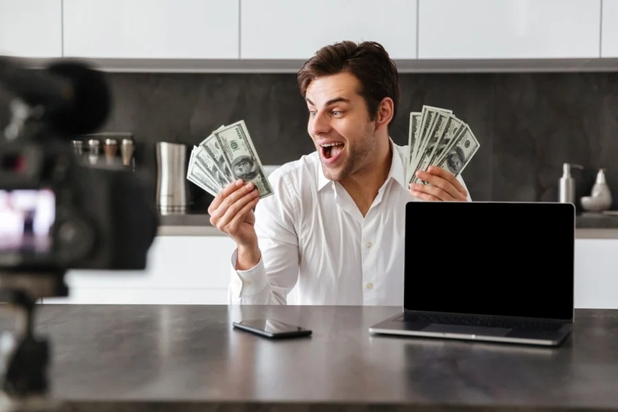 Homem faz expressão de surpresa agradável enquanto segura maços de dinheiro em ambas as mãos. Ele está em frente à uma câmera. Há um notebook e um celular na mesa à sua frente. Representando como ganhar dinheiro pela internet.