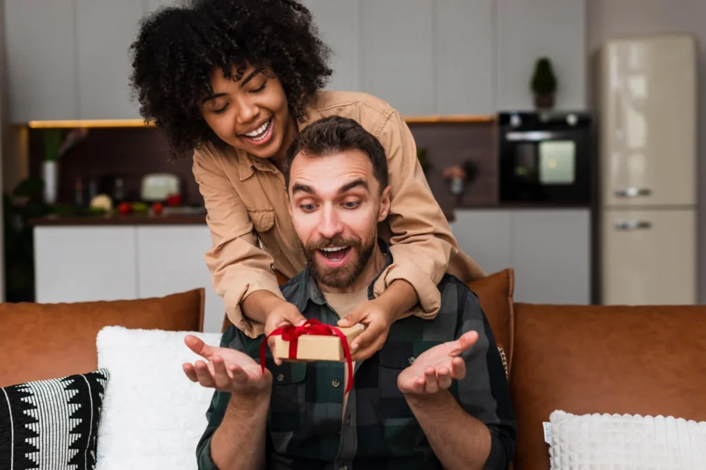 Casal sorridente em casa; mulher surpreende o rapaz com um gift card Tesouro Direto em caixa vermelha.