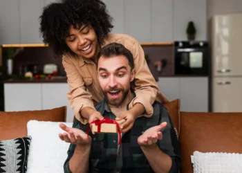 Casal sorridente em casa; mulher surpreende o rapaz com um gift card Tesouro Direto em caixa vermelha.