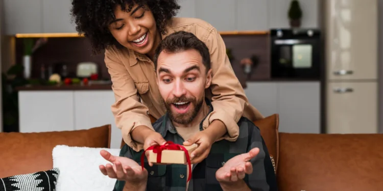 Casal sorridente em casa; mulher surpreende o rapaz com um gift card Tesouro Direto em caixa vermelha.