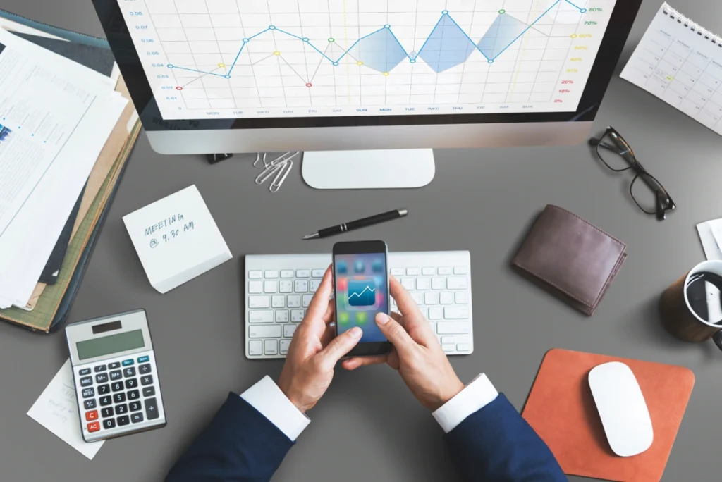 Homem mexe em app de finanças em sua mesa de trabalho, que também tem um computador, uma calculadora, uma carteira e outros elementos. Falando sobre os melhores aplicativos de finanças.