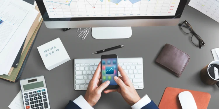 Homem mexe em app de finanças em sua mesa de trabalho, que também tem um computador, uma calculadora, uma carteira e outros elementos. Falando sobre os melhores aplicativos de finanças.