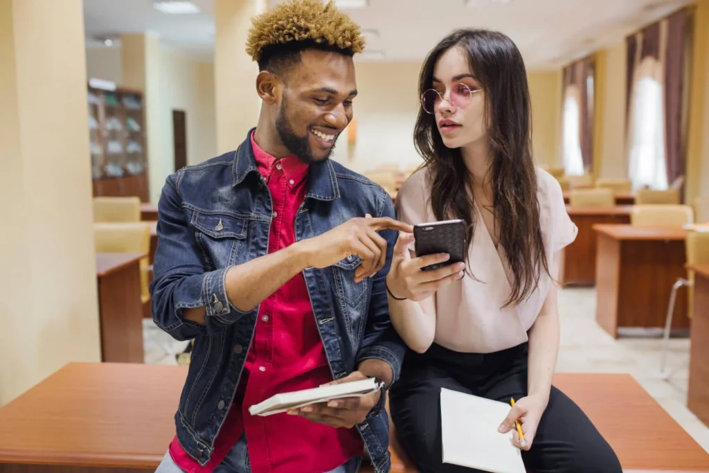 Dois jovens em ambiente acadêmico olhando para celular, representando cartões de crédito sem anuidade.