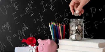 Finanças pessoais: organize seu orçamento hoje de forma prática 3 Pessoa coloca moeda em cofre improvisado em um copo de vidro. Há uma pilha de livros, um cofre em formato de porquinho e outros elementos na imagem. Representando "organizar orçamento".