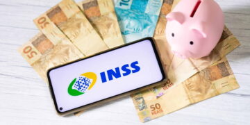 INSS divulga datas de pagamento de março; confira o calendário completo 1 Celular com símbolo do INSS na tela sobre notas de real espalhadas. Há um cofre em formato de porquinho na imagem, representando pagamento do INSS.