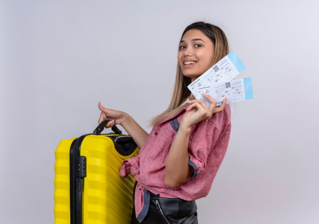 Mulher sorridente exibe bilhetes de passagens aéreas enquanto segura uma mala de viagem amarela, representando passagens aéreas baratas.