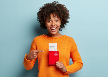 Mulher sorridente segura e aponta para passaporte junto com passagem aérea.