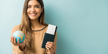 Descubra os passaportes mais valiosos do mundo em 2025 3 Mulher exibe um dos passaportes mais valiosos do mundo em uma das mãos e segura um pequeno mapa em forma de globo Terra na outra mão.
