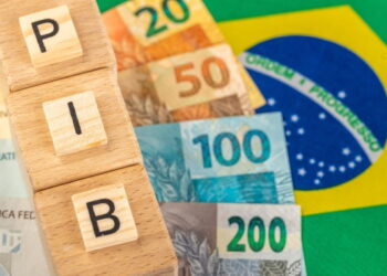 Bandeira do Brasil ao fundo, notas de real e cubos formando o nome "PIB" em primeiro plano.
