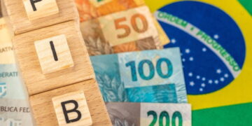 Bandeira do Brasil ao fundo, notas de real e cubos formando o nome "PIB" em primeiro plano.