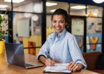 Empreendedora sorri para a foto, enquanto analisa gráficos em seu notebook. Falando sobre programas do governo para empresas.