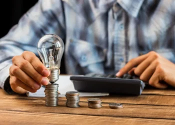 Homem usa uma calculadora ao mesmo tempo que posiciona uma lâmpada sobre pilhas de moedas que estão sobre uma mesa, representando tesouro rende mais.