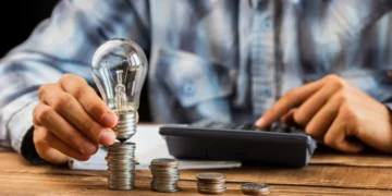 Tesouro Renda Mais: o que é, como funciona e como começar a investir 5 Homem usa uma calculadora ao mesmo tempo que posiciona uma lâmpada sobre pilhas de moedas que estão sobre uma mesa, representando tesouro rende mais.