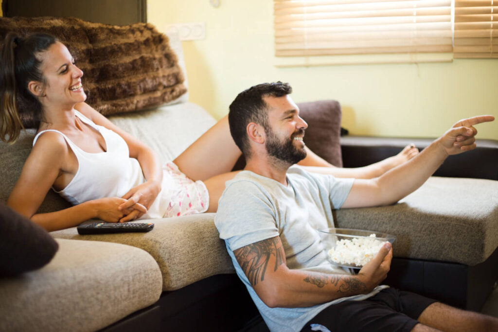 Casal relaxa no sofá assistindo séries em alta na Netflix, sorrindo e comendo pipoca.