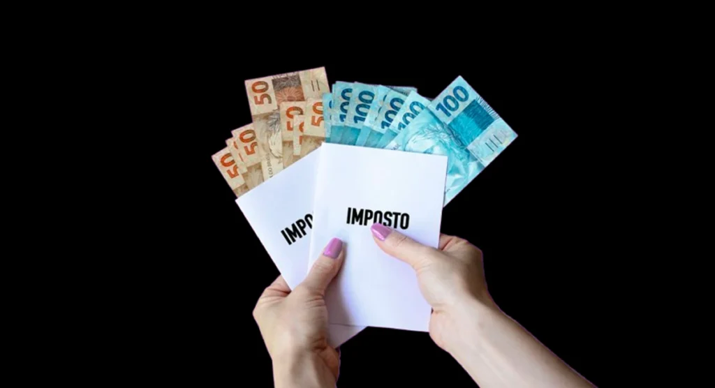 Mãos de uma mulher segura notas de R$ 50 e R$ 100 e dois folhetos onde está escrito "imposto". Falando sobre a tributação no Brasil.