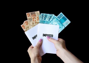 Mãos de uma mulher segura notas de R$ 50 e R$ 100 e dois folhetos onde está escrito "imposto". Falando sobre a tributação no Brasil.