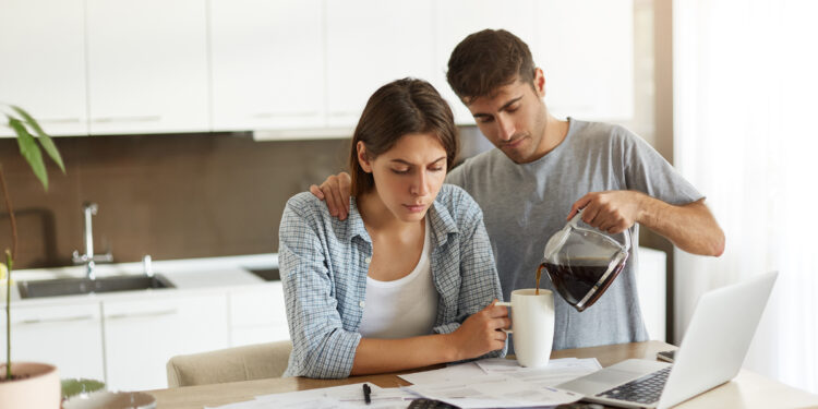 Picture of young male and female doing paperwork together at home: serious wife sitting at dining table with papers and laptop computer, calculating bills while her husband serving coffee to her